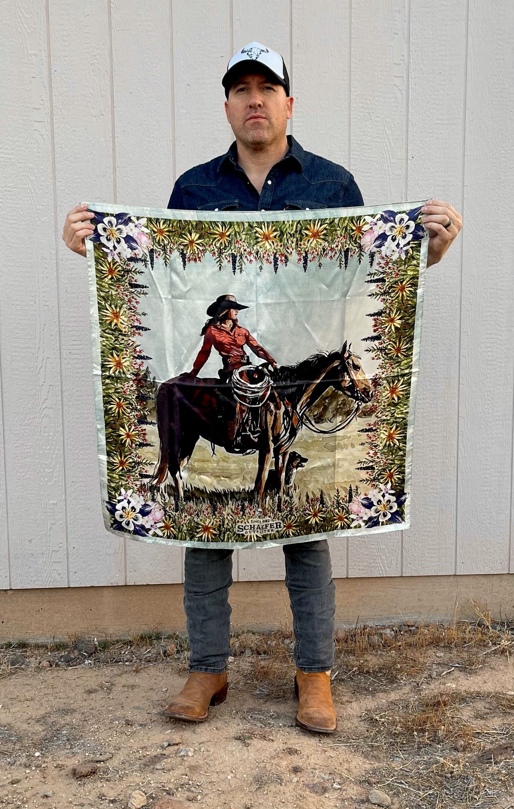 Wild rag scarf held up outdoors, featuring cowboy on horse with colorful floral edge.