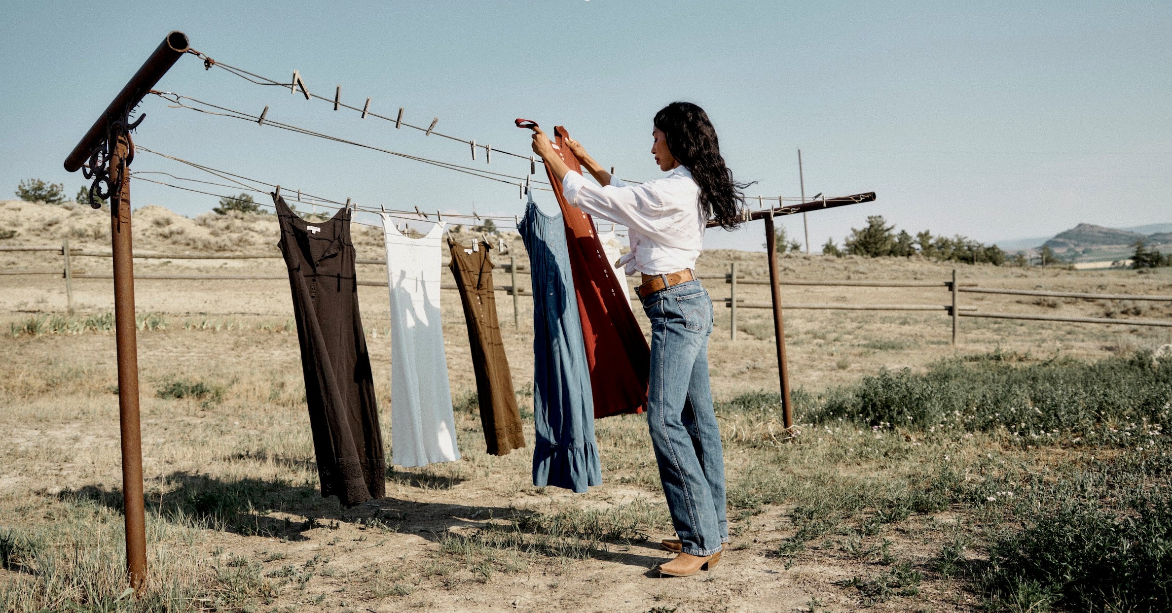 A woman in a white long-sleeve top and blue jeans hangs a rust-colored dress on a rustic metal clothesline in an open grassy field under a clear blue sky.